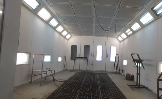 The image shows an interior view of a workshop with white walls and a clean environment, featuring a large metal table at the center, surrounded by various tools and equipment, including a welding machine, benches, and other machinery typically found in a workshop setting.