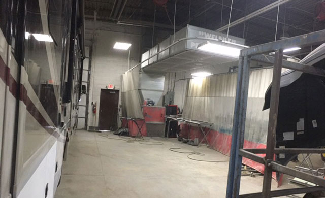 The image shows an interior space with a large recreational vehicle RV parked inside a building, featuring a spacious living area with a couch and a television screen, a kitchenette with a countertop and sink, and a dining table with chairs.