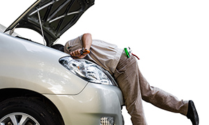 The image shows a person working on the engine of a silver car with the hood open, standing next to it with tools and wearing a uniform.