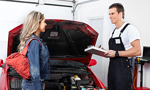 The image shows a man and woman standing next to an open car hood in a garage setting.