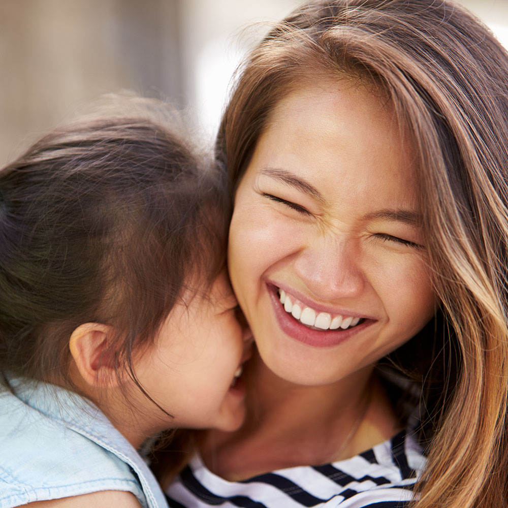 A woman with long dark hair is smiling at her child who is laughing while being held by the woman. They are both outdoors, possibly on a sunny day, given the bright lighting in the photo.